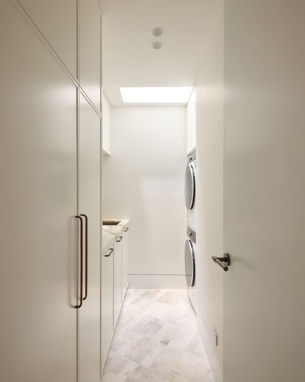 A narrow, bright laundry room viewed through an open doorway. Full-height white cabinetry with long dark handles lines the left side, with a benchtop and sink area beyond. Stacked front-loading washer and dryer units are built into the right wall. The room has white walls and ceiling, a rectangular skylight, recessed downlights, and pale patterned tile flooring.
