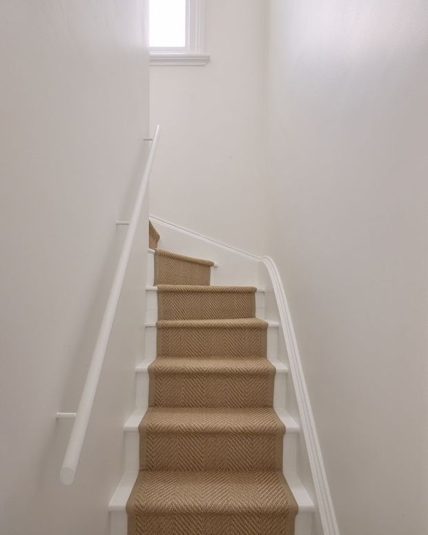 A narrow residential staircase viewed from below, with a beige patterned carpet runner, white painted stair risers and trim, white walls, and simple white handrails. A small upper window provides soft natural light, creating a clean and understated traditional interior.