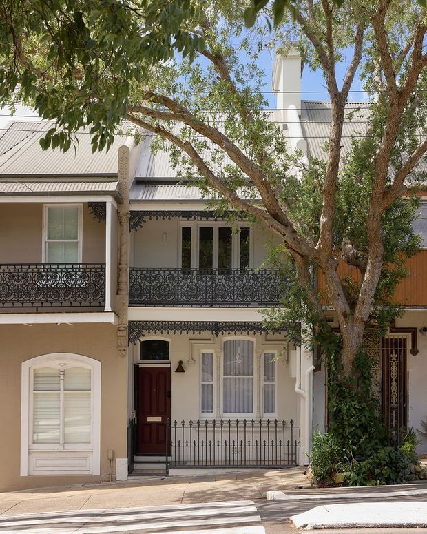 Street-facing view of a narrow, attached two-storey terrace or townhouse with a pale rendered facade, corrugated metal pitched roof, prominent chimney, decorative wrought-iron balcony balustrades, iron window and door grilles, and traditional ornamental trim. Mature trees partly frame the facade and cast dappled shade across the footpath.