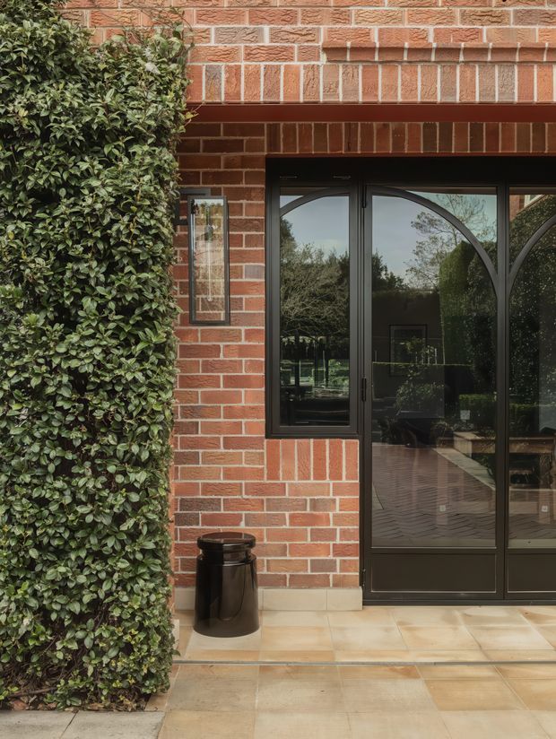 Close-up exterior view of a brick building entrance with dark-framed glazed double doors and a narrow side window. The facade features red-orange brickwork with decorative projecting brick bands and a pale stone or concrete threshold. A dense clipped hedge borders the entrance, which opens onto a paved terrace or walkway. Reflections in the glazing show landscaped outdoor surroundings.