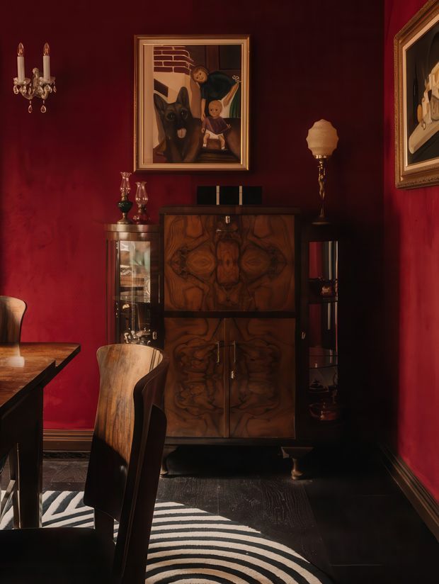 A richly styled, moody dining room corner with deep red walls, dark flooring, a sculptural timber dining table and chairs, and an ornate dark wood cabinet. Decorative glassware, framed artwork, a vintage-style lamp, and a striped rug create a dramatic, eclectic atmosphere with strong warm lighting.