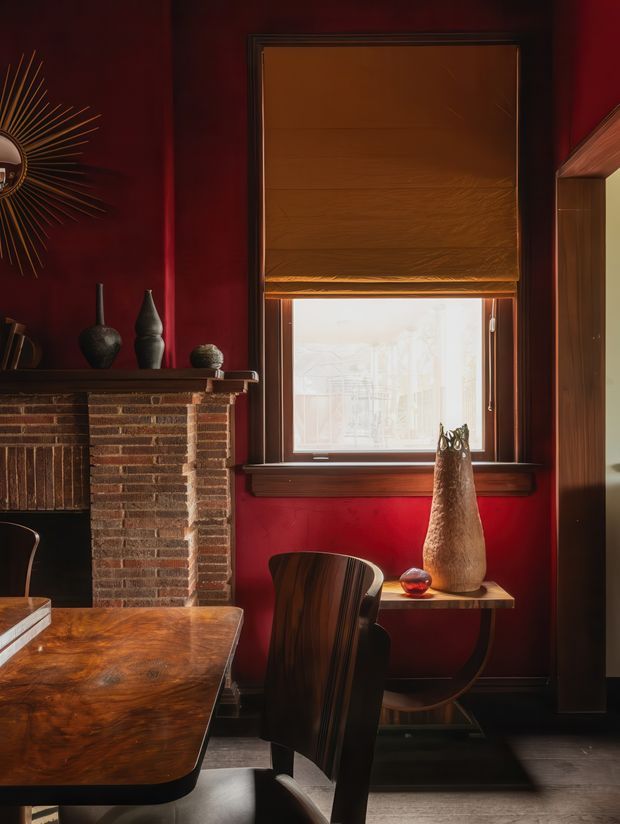 A richly styled, moody interior corner featuring deep red walls, a dark brick fireplace with timber mantel, a wood-framed window with a brown roller blind, and dark timber furniture. A sculptural sunburst mirror, ceramic vessels, a tall textured vase, and a polished timber dining table add warm decorative detail. The room appears to function as a dining or living area, with a doorway or opening visible at right.