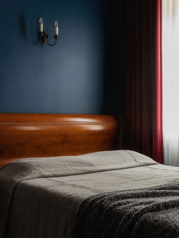A moody bedroom interior centered on a bed with layered grey bedding and a dark textured throw. The bed has a substantial polished dark timber headboard with traditional curved detailing. A deep blue painted wall features a small black double-arm wall sconce, while dark red curtains and sheer white window coverings bring in soft daylight from the right.