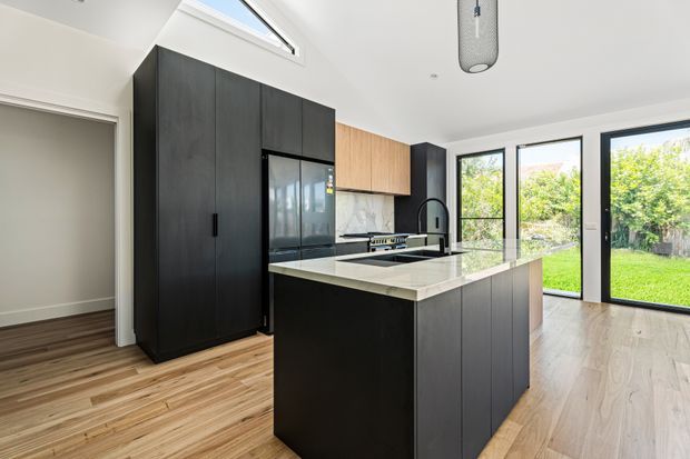 A bright contemporary kitchen featuring a large dark timber-toned island with a pale stone benchtop, integrated black cabinetry and appliances, warm timber wall cabinetry, timber flooring, and extensive black-framed glazing opening to a green garden. A skylight adds additional natural light.