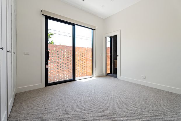 Bright, empty bedroom with neutral painted walls, grey textured carpet, built-in white wardrobe cabinetry, and black-framed floor-to-ceiling glazed doors opening to a small brick-enclosed outdoor area. A secondary internal doorway is visible on the right.