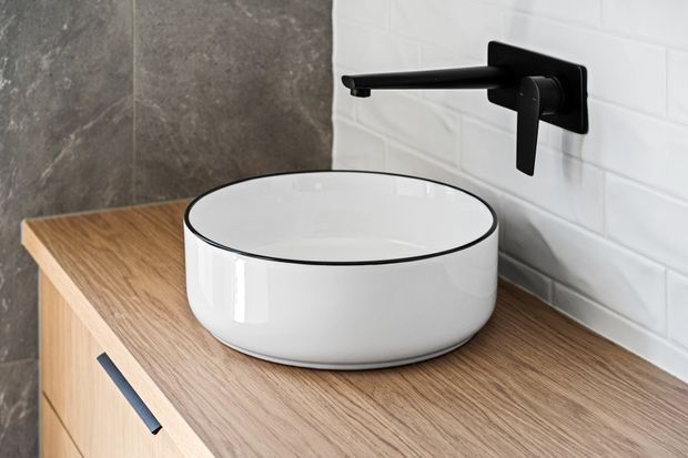 Close-up of a contemporary bathroom vanity featuring a white round vessel basin with a thin black rim, matte black wall-mounted tapware, and a timber-look benchtop with matching cabinetry. The wall combines dark grey stone-look tiling with white subway tiles, creating a refined high-contrast finish.