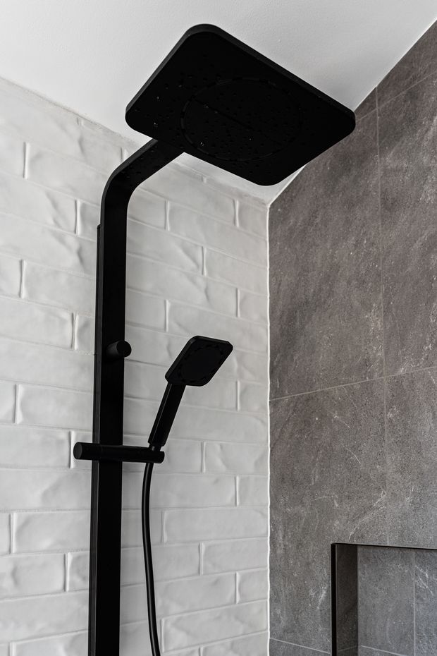 Close-up of a modern black matte shower system with a large square overhead rain shower and handheld shower mounted against contrasting white textured subway tile and dark grey stone-look wall tile. A recessed wall niche is partially visible at the lower right.
