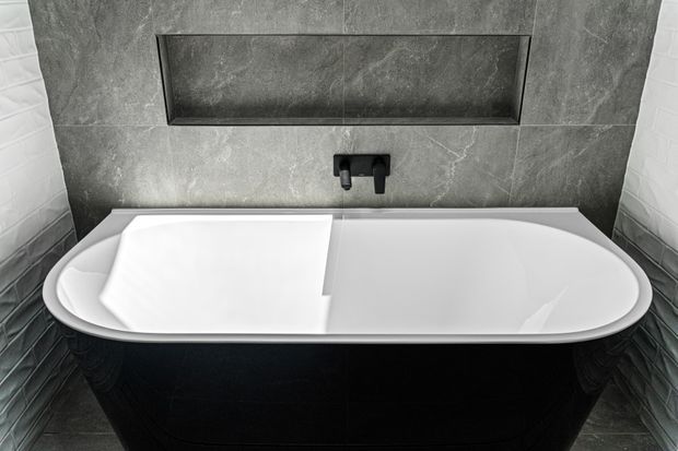 Close-up view of a freestanding white bathtub with a black exterior, set against dark grey stone-look wall tiles. A recessed wall niche and matte black wall-mounted tapware are visible above the bath, with matching dark floor tiles.