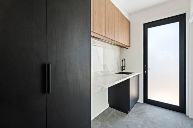Compact contemporary laundry with full-height matte black storage cabinetry, warm timber overhead cupboards, a white marble-look splashback and benchtop, black undermount sink and tapware, and grey tiled flooring. A black-framed frosted-glass exterior door provides natural light.