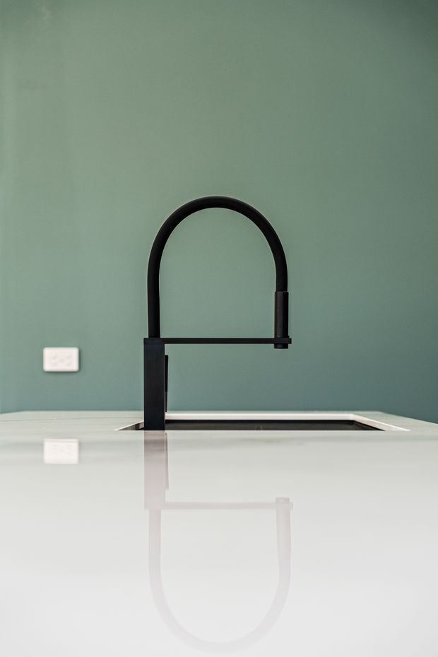 Close-up view of a minimalist kitchen sink area featuring a tall matte black gooseneck tap mounted through a white countertop, with a dark inset sink and a muted sage-green painted wall. The reflective countertop creates a soft mirror image of the tap and nearby wall outlet.
