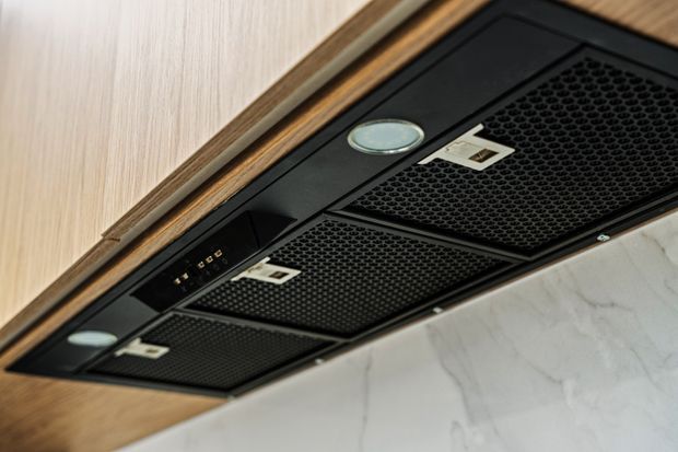 Close-up view of a sleek black integrated rangehood beneath light timber cabinetry, with multiple honeycomb-patterned filters, recessed controls, and small built-in lights. A pale marble-look wall surface is visible behind it.