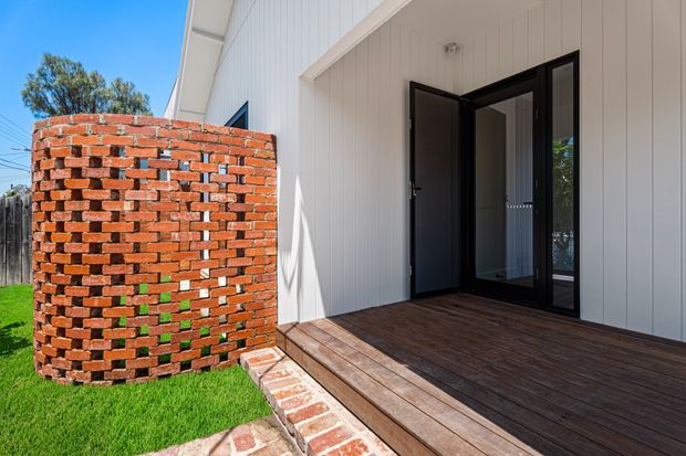 Exterior view of a contemporary white-clad building entrance with a dark timber deck, black-framed glazed double doors, and a curved exposed-brick privacy wall beside a small lawn.