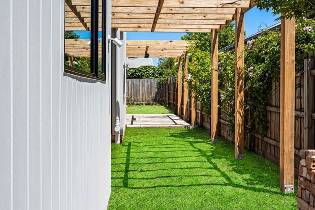 A narrow landscaped side passage beside a white-clad building, featuring a timber pergola with repeated overhead beams, a bright green lawn, timber fencing, and a small timber deck at the far end. Strong sunlight casts geometric pergola shadows across the grass.