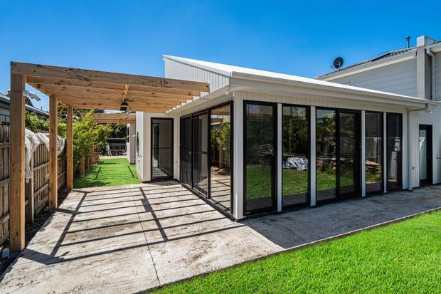 Exterior view of a contemporary single-storey home extension with a covered timber pergola over a concrete patio and walkway. The extension has pale vertical weatherboard cladding, a low sloping metal roof, and extensive black-framed glazing and doors opening toward the garden. Bright suburban outdoor setting with lawn and timber fencing.