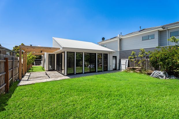 Sunny exterior view of a contemporary single-storey extension or garden room with extensive black-framed glazing, a light metal pitched roof, and a timber pergola over a paved patio. The building sits beside a larger light-grey weatherboard-clad house, with a broad green lawn and timber fencing in the foreground.