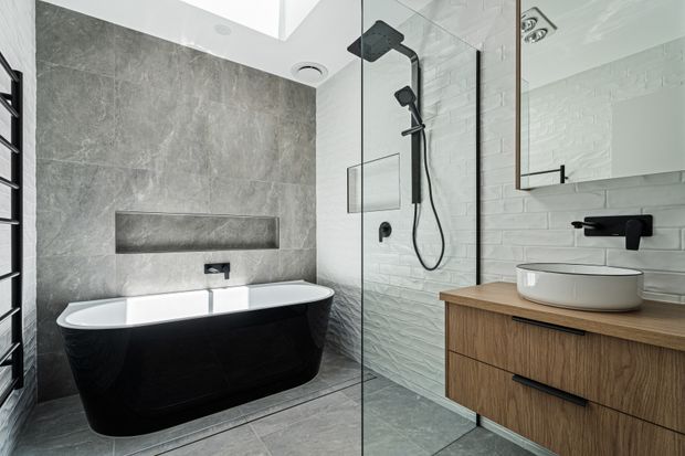 Contemporary bathroom featuring a freestanding black bathtub against a large grey stone-look feature wall, a walk-in glass shower with black fixtures, white textured wall tiles, and a timber vanity with a white vessel basin. A skylight and recessed ceiling lighting create a bright, refined atmosphere.