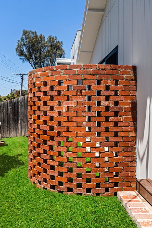 Exterior view of a contemporary home featuring a curved, perforated red brick privacy wall beside a white vertically clad facade. The brickwork incorporates irregular openings and patterned gaps, with a small brick-paved threshold, timber decking, lawn, fencing, and a mature tree visible around the building.