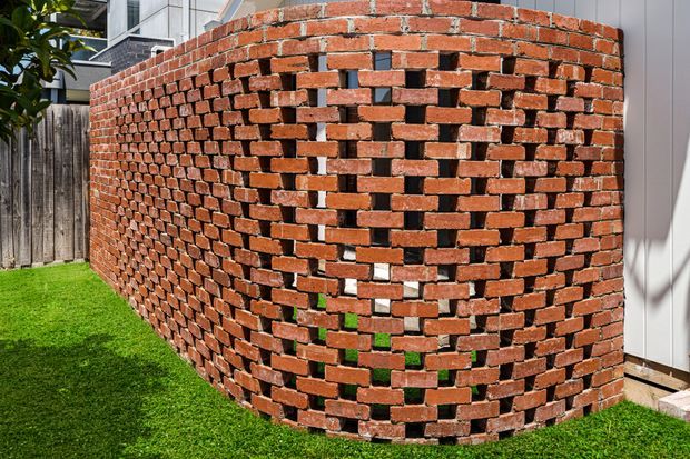 Exterior view of a curved red brick masonry wall constructed with an open, perforated bond pattern. The wall forms a sculptural privacy screen beside a grass lawn, with timber fencing and part of a contemporary building visible behind it.