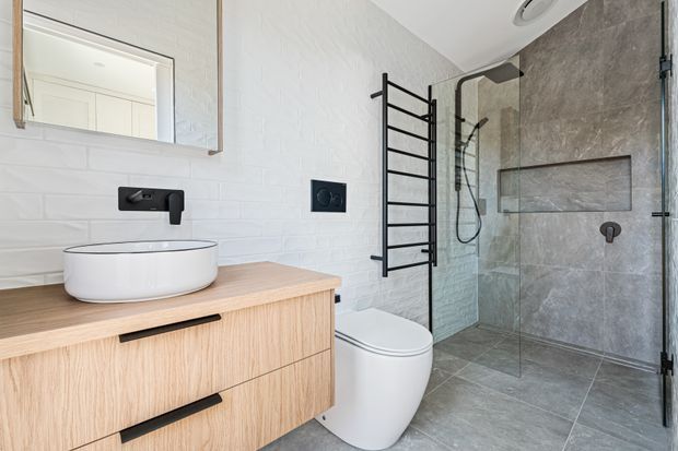 Contemporary bathroom featuring a light timber floating vanity with a white vessel basin, matte black tapware and hardware, white brick-pattern wall tiles, a wall-hung toilet, black heated towel rail, and a frameless glass walk-in shower with large grey stone-look tiles.