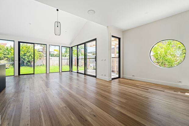 Bright, largely unfurnished open-plan interior with white walls and ceiling, extensive black-framed glazed doors and windows opening toward a green outdoor area, warm timber flooring, a distinctive oval window, and a simple hanging pendant light.