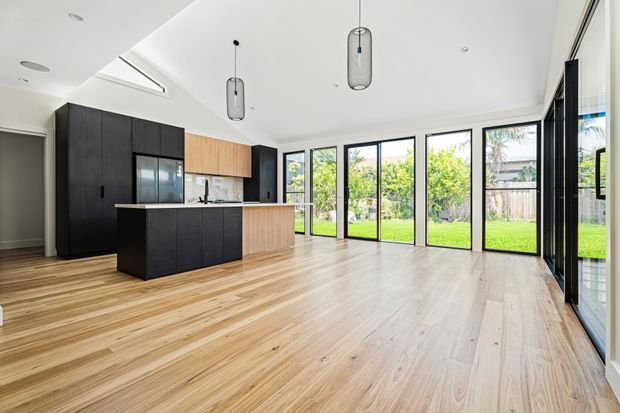 Bright open-plan kitchen and living area with extensive floor-to-ceiling black-framed glazing, pale timber flooring, and a contemporary kitchen featuring dark cabinetry, timber upper cabinets, and a central island. Pendant lights hang from a high white ceiling, with a leafy garden visible outside.