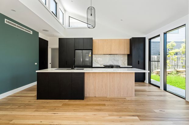 A bright contemporary kitchen with a large central island, dark cabinetry, light timber cabinetry, a white stone benchtop, and a marble-look splashback. The room has warm timber flooring, a dark green feature wall, full-height glazing and a high ceiling with skylights. A garden is visible through the glazed doors.