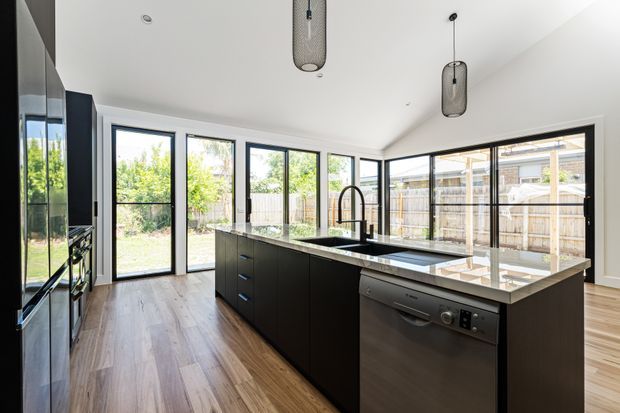 A bright contemporary kitchen with a large central island, dark built-in cabinetry, pale stone benchtops, integrated appliances, and black-framed floor-to-ceiling glazing opening toward a fenced garden. Warm timber flooring and pendant lights add contrast to the predominantly white and charcoal palette.