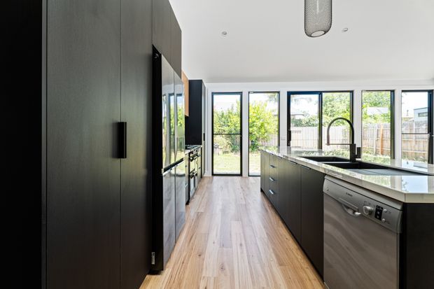 A contemporary galley-style kitchen with a large central island, dark flat-panel cabinetry, a pale stone benchtop, integrated appliances, and a stainless-steel dishwasher. Warm timber flooring contrasts with the black joinery, while full-height windows and glazed doors provide abundant natural light and views to the garden.