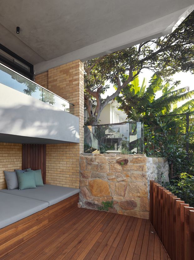 A compact outdoor terrace or balcony with a warm timber deck, built-in grey upholstered seating beneath a concrete overhang, exposed tan brick walls, a frameless glass balustrade, and a rustic natural-stone retaining wall. Dense tropical greenery surrounds the space, creating a sheltered, leafy outdoor setting.