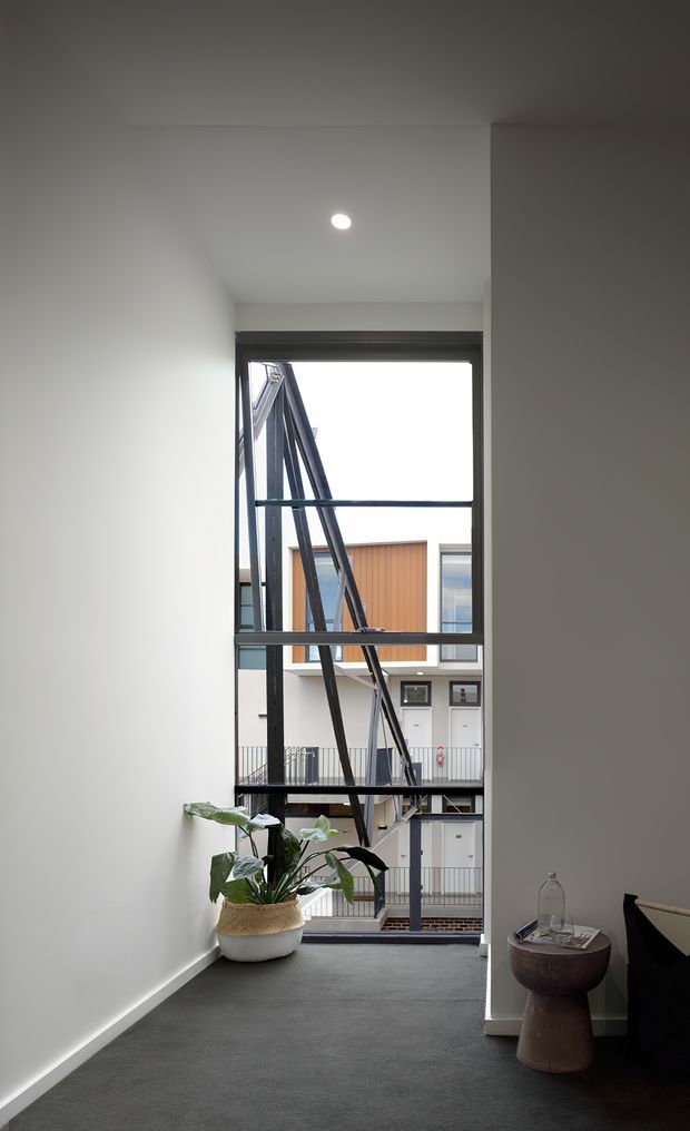 A narrow, contemporary interior landing or hallway with white walls, dark carpet, and a high floor-to-ceiling window overlooking an internal multi-level courtyard or atrium. The black-framed glazing and exposed external stair structure create a strong architectural feature. A potted plant and small sculptural side table sit beside dark upholstered seating. The space is softly lit by a recessed ceiling light and natural daylight.