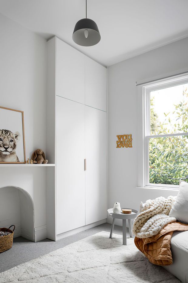 Bright, softly styled bedroom corner with a tall white built-in wardrobe, open shelving, arched alcove, framed animal artwork, pendant light, window, small side table, and bed with layered knitted and quilted bedding. The room uses a restrained white and light-grey palette with warm timber and tan accents.