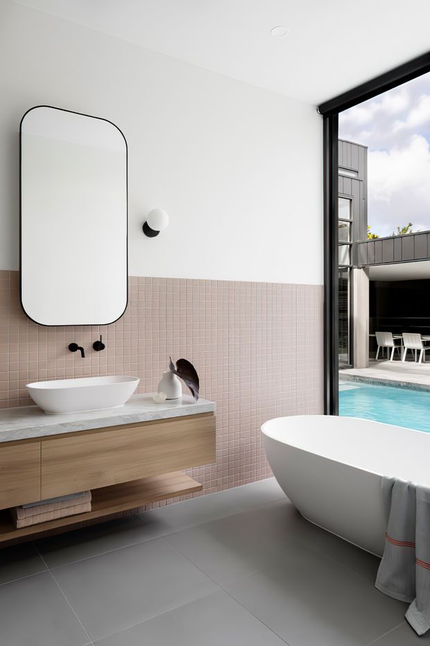 Contemporary bathroom with a freestanding white bathtub, wall-mounted timber vanity, vessel basin, large rounded-corner mirror and minimalist black wall light. The lower walls are clad in pale pink square tiles, while large grey floor tiles create a calm, refined palette. Full-height black-framed glazing opens views to an outdoor pool and adjacent dining terrace.