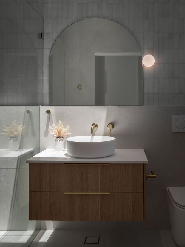 A moody contemporary bathroom featuring grey tiled walls, a large arched recessed wall detail, wall-mounted round globe lighting, and a floating dark timber vanity with a pale stone benchtop and white vessel basin. Brass-toned tapware and handles add warmth, while a glass shower screen and toilet are visible.
