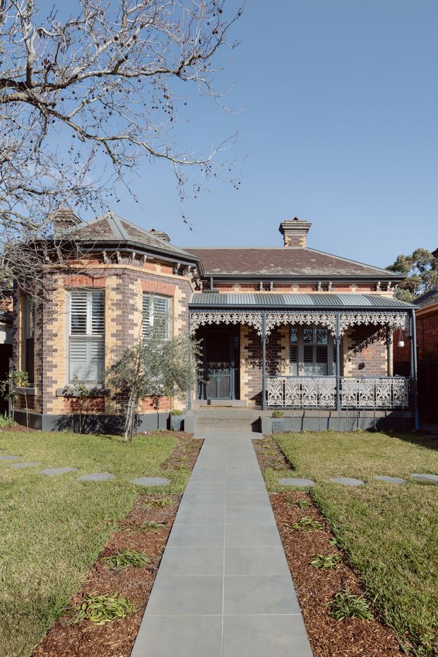 Front exterior view of a single-storey heritage brick house with an elaborate lacework veranda, decorative brick detailing, timber-framed windows with shutters, a hipped roof, and a central chimney. A straight paved path leads through the front lawn to the veranda.