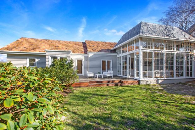 Exterior view of a single-storey light-grey weatherboard home with a terracotta tiled pitched roof and a prominent glazed sunroom or conservatory extension. A timber deck with outdoor seating connects the house to the landscaped lawn and garden.