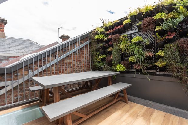 A rooftop terrace with timber decking, built-in picnic-style dining benches and table, and a large vertical garden mounted against a dark wall. A metal balustrade overlooks adjoining brick buildings and slate roofs. The space combines urban outdoor dining with lush planted screening.