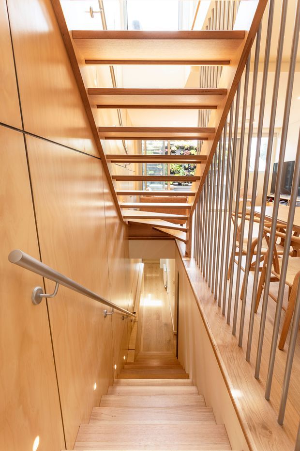 Bright interior view looking down a timber-lined stairway. The staircase has pale timber treads and wall panelling, a stainless-steel handrail, and a slim vertical metal balustrade. Open timber beams overhead create a partially open stairwell, with natural light entering through large glazing above and a glimpse of dining furniture to the side.