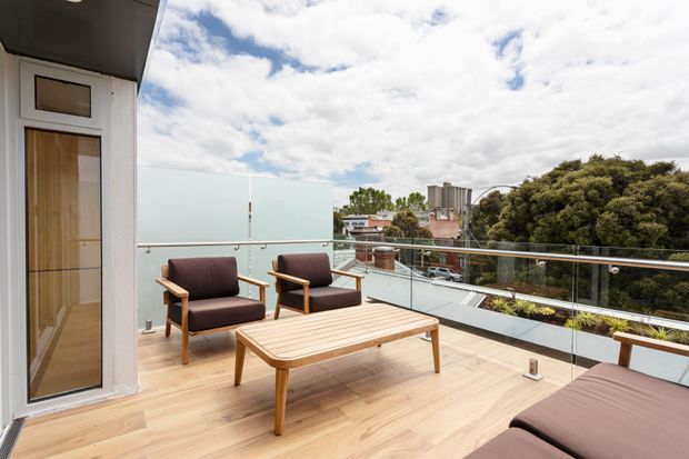 A contemporary rooftop terrace or balcony with light timber decking, glass balustrades, and a relaxed outdoor seating arrangement. Two timber-framed armchairs and a low slatted coffee table face the view, with part of a cushioned lounge visible in the foreground. A glazed door connects the terrace to the interior, and the urban residential surroundings are visible beyond.