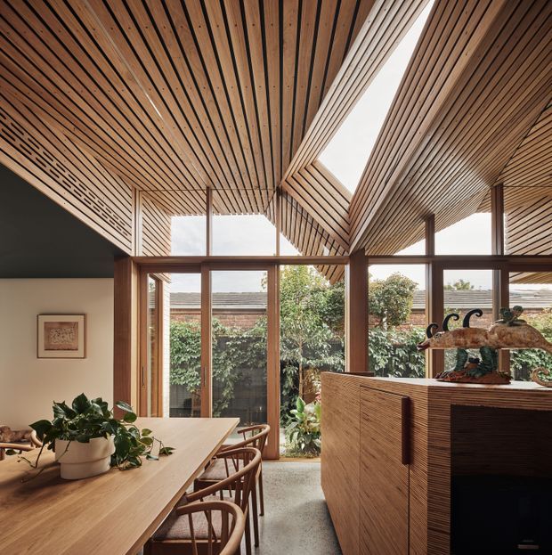 A warm, architecturally detailed dining and living area with a long timber dining table, curved timber dining chairs, indoor plants, and a timber-clad fireplace or joinery unit. Full-height glazing opens views to a planted courtyard. The defining feature is the richly expressed timber slatted ceiling, which folds around a large linear skylight and is supported by timber-framed glazing and posts.