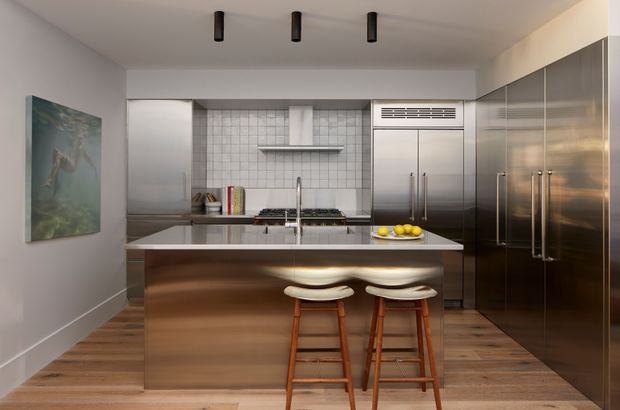 Contemporary galley-style kitchen with a large central island and two timber bar stools. The space features dark stainless-steel appliances and tall cabinetry, a white square-tile splashback, stainless-steel rangehood and sink, warm timber flooring, white walls, and black cylindrical ceiling downlights. A large artwork hangs on the left wall.