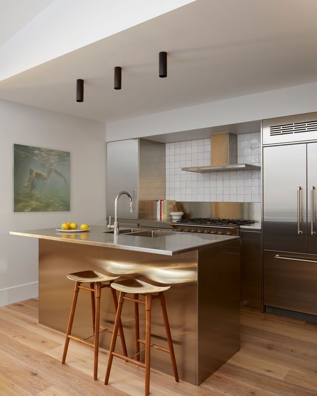 Contemporary kitchen with a large island, integrated stainless-steel appliances, metallic cabinetry, white tiled splashback, timber flooring, and sculptural bar stools. Recessed ceiling spotlights and a restrained neutral palette create a refined, modern interior.
