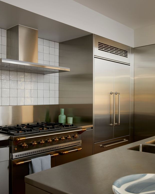 A refined commercial-style kitchen with stainless-steel appliances and cabinetry, a large professional gas range, tiled splashback, overhead extraction canopy, and a broad stone island benchtop. Brass-toned hardware and dark metallic surfaces add warmth and contrast to the restrained neutral palette.