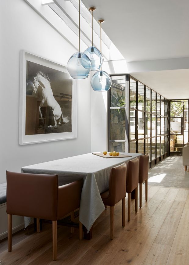 Bright, refined dining room with a long rectangular table covered in a light linen cloth and surrounded by warm brown upholstered dining chairs. A cluster of blue glass pendant lights hangs above the table, with a large framed monochrome horse artwork on the wall. Skylights and a glazed, black-framed partition bring abundant natural light into the adjoining space. The room has wide light timber flooring and a calm contemporary feel.