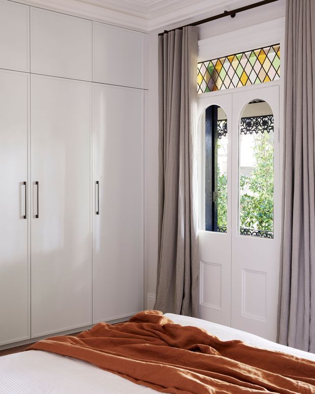 Bright, refined bedroom featuring full-height white built-in wardrobes, a white bed with rust-coloured bedding, and a tall glazed French-style door with decorative leadlight and black wrought-iron detailing. Soft taupe curtains frame the door, while ornate ceiling cornices and panelled joinery add classic architectural character.