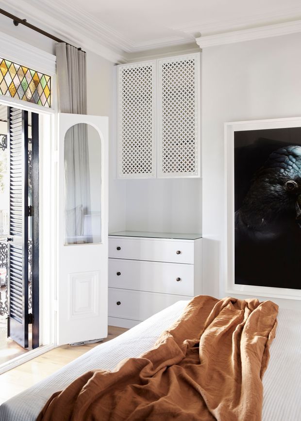 Bright, refined bedroom with white built-in cabinetry, a glass-topped drawer unit, decorative lattice-front overhead cupboards, ornate ceiling cornices, and a tall white door with an arched inset mirror. A stained-glass transom and open louvred security door bring in natural light. The bed is dressed in white linen with a rust-brown throw, and a large framed dark bird artwork forms a dramatic focal point.