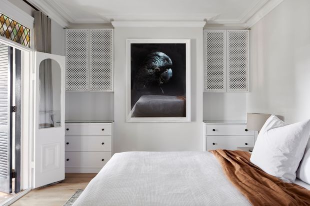 Bright, refined bedroom with a white upholstered bed, layered white bedding, and a warm brown throw. Built-in white cabinetry and drawer units flank a central framed artwork, while decorative lattice-front storage panels, ornate ceiling moulding, timber flooring, and a glazed timber door add character. Natural light enters through the open door and louvred glazing.