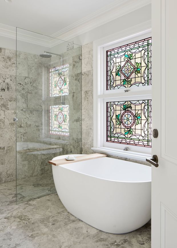Bright, refined bathroom featuring a freestanding white bathtub with a timber bath tray, a frameless glass walk-in shower, and pale natural stone wall and floor tiles. Decorative stained-glass windows with botanical and geometric motifs provide a colourful heritage-style focal point, complemented by white panelled detailing and crown moulding.