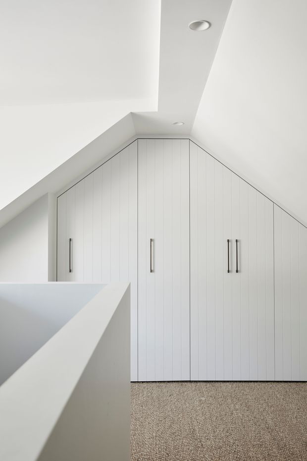 A minimalist upper-level storage area with full-height white built-in cabinetry fitted beneath a sloping roof. The cabinet doors have subtle vertical panelling and slim dark metal handles, while a white balustrade borders the foreground stair opening. The room has white walls and ceiling, recessed downlights, and a neutral textured carpet.