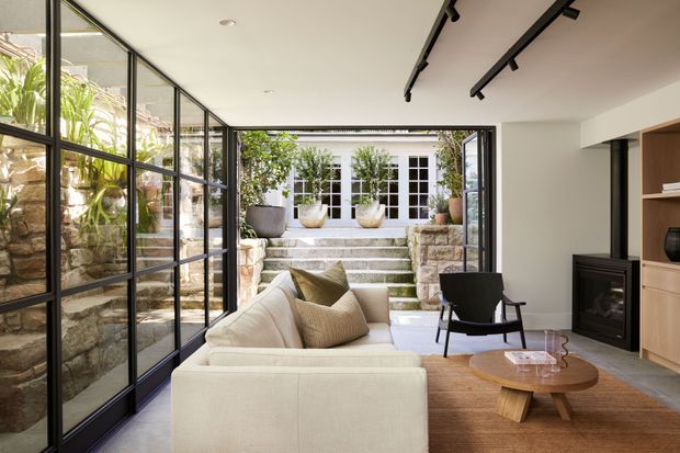 A refined contemporary living room opening onto a planted stone courtyard. The room features floor-to-ceiling black-framed glazing, a neutral upholstered sofa, black lounge chair, timber coffee table, warm timber flooring, built-in timber shelving, and a slimline fireplace. Stone steps and garden planting are visible beyond the fully opened glazed doors.