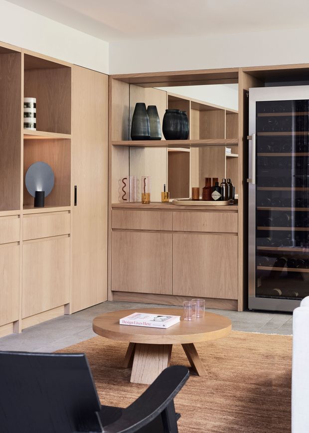 A warm, refined living room with extensive light timber built-in cabinetry, open shelving, mirrored backing, and a large integrated glass-front wine refrigerator. The room includes a round timber coffee table, neutral rug, sculptural decor, and dark lounge seating.